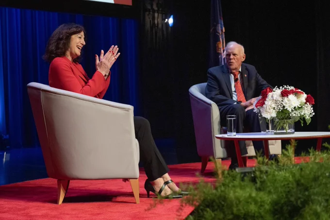 President Goldsmith and John Hennessy are seated on a stage are engaged in conversation. President Goldsmith is clapping. A table with a floral arrangement and two glasses of water is placed between them. 
