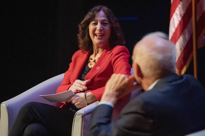 President Goldsmith is smiling and holding papers. John Hennessy is seated nearby, facing them. An American flag is visible in the background.
