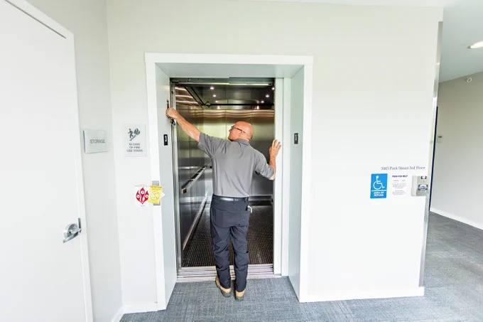A person is holding the door of a silver Otis elevator open in a modern building. There are accessibility symbols on the wall near the elevator entrance and a sign indicating "Stairwell" beside a nearby door.