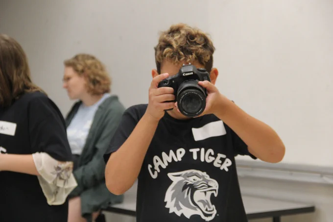 A child uses a camera while wearing a shirt that reads: Camp Tiger 