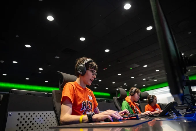 Children wearing gaming headsets sit in a large computer lab playing computer games 
