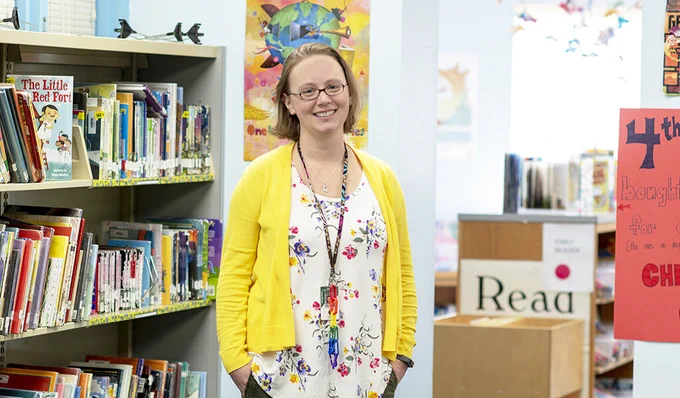 A librarian stands and smiles in front of book shelves at a school library 