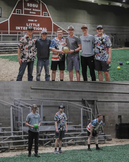 Top photo: Participants stand together on the wood chips inside the SDSU Animal Science Arena during an Ag Week dodgeball event, holding a dodgeball for a group photo.
Bottom photo: Participants get ready to start playing Dodgeball in the SDSU Animal Science Arena. 