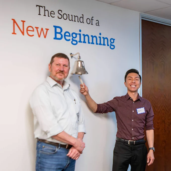 Two men stand by a wall with the phrase 'The Sound of a New Beginning,' one ringing a bell symbolizing a fresh start.
