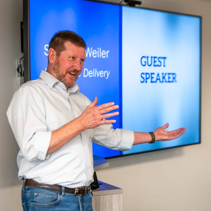 Man in casual attire giving a presentation with a screen behind him displaying 'Guest Speaker'.