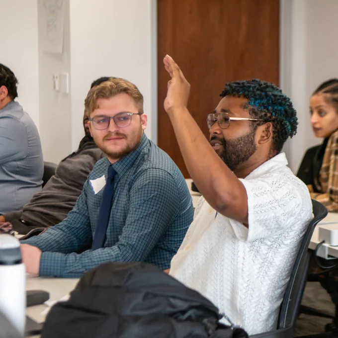 A group of diverse adults seated in a classroom setting, one man raising his hand to ask a question during a discussion.