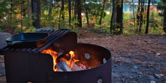 a steel fire pit with a fire lit inside it and a cast iron pan on the grill part of the fire pit.