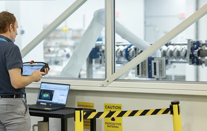 A person operates a control panel next to a laptop in a laboratory setting. The lab is separated by a glass wall, behind which robotic arms are visible. There are caution signs indicating radioactive material.