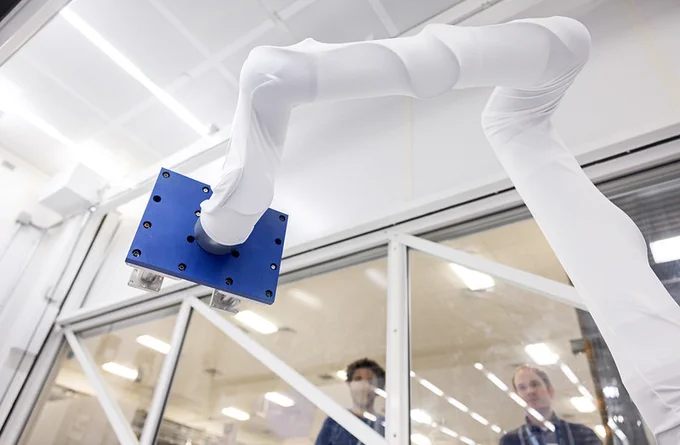 A large white robotic arm inside a clear-walled room is handling a flat, blue rectangular board. Two people observe the robot's operation from behind a window. 