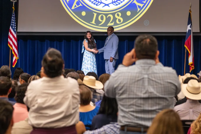 A woman and a man shake hands on a stage. A seated audience watches, with some people taking photos.