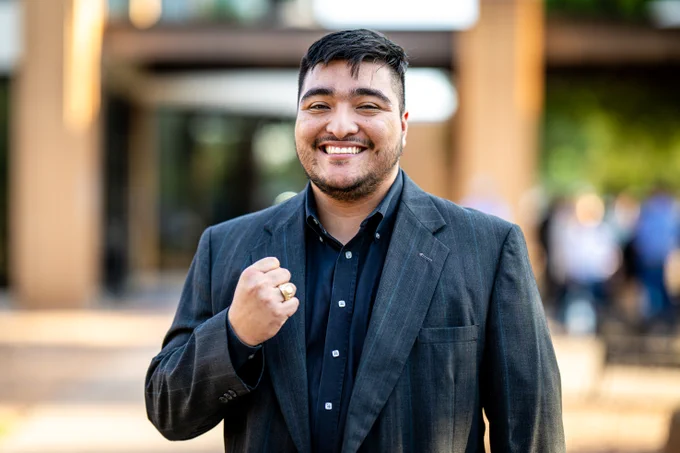 A smiling man stands outdoors, making a fist with his right hand. He wears a ring on his right ring finger.