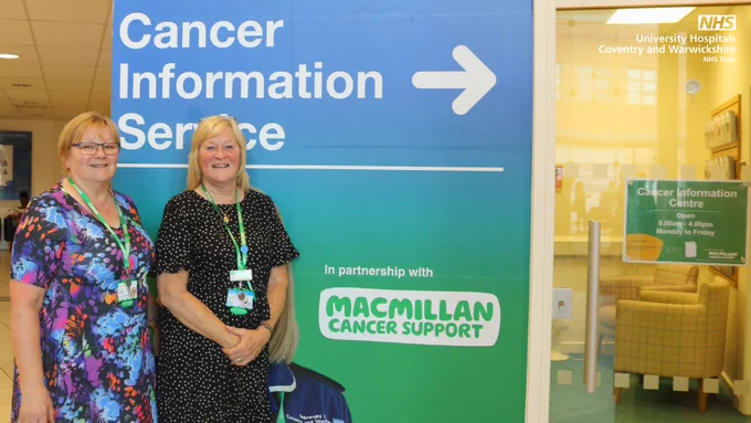 Two staff members stand beside a large blue and green sign reading “Cancer Information Service →” at University Hospitals Coventry and Warwickshire NHS Trust, in partnership with Macmillan Cancer Support. The sign is located near the hospital’s main entrance, with the Cancer Information Centre visible through glass doors in the background.