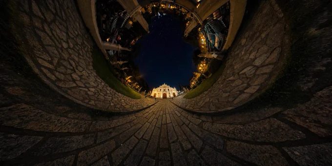 Reversed tiny planet projection from a 360° panorama of Vigan Church. Curved cobblestone pathways wrap inward toward a warmly lit historic church at night, with surrounding buildings and trees forming a circular horizon. No stars in the sky, emphasizing the soft glow and shine of the church as the focal point.