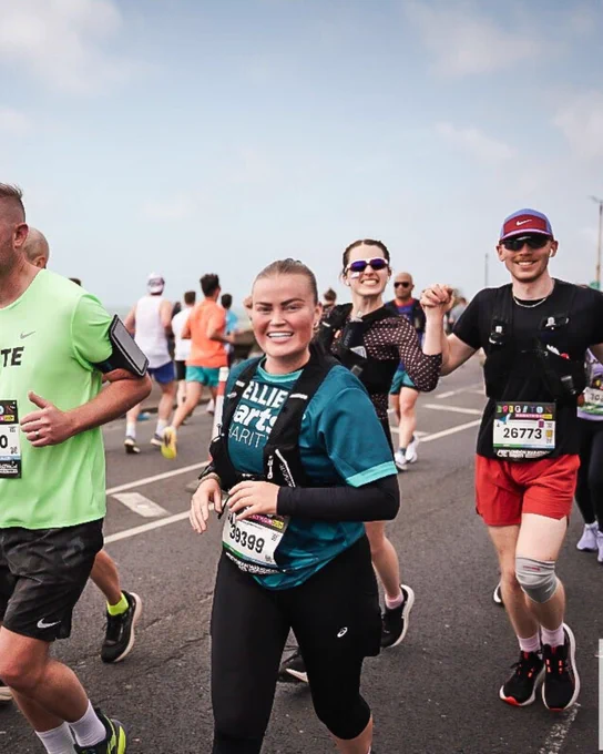 Runners participating in a marathon, smiling and holding hands, with several wearing numbered bibs and athletic gear. Others are visible running in the background on a road.