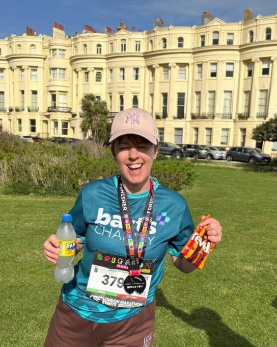 A person is standing outdoors, holding a bottle of Fanta and a pack of sweets. They are wearing a marathon finisher medal and a "Barts charity shirt. A large, elegant building with multiple windows is in the background.