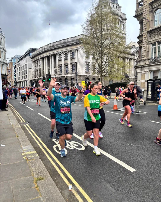 A group of marathon runners in colorful athletic wear are participating in a street race. Some runners are wearing “NSPCC” shirts. Spectators line the sidewalk, and historic buildings are visible in the background. One runner waves cheerfully at the camera.