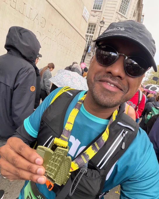 A person wearing sunglasses and a cap holds up a medal in front of the Smithsonian National Gallery. The background includes several people, some with umbrellas.