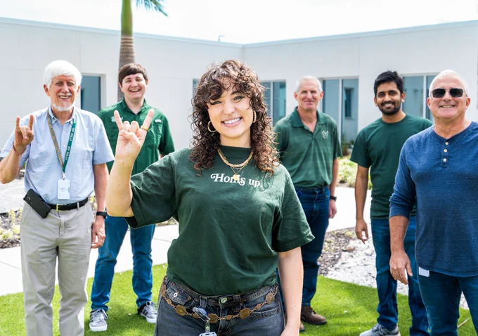 A group of six people standing outside a building on a grassy area. One person in front wears a "Horns up!" shirt, and the others are behind, some making the "Horns Up" hand symbol.