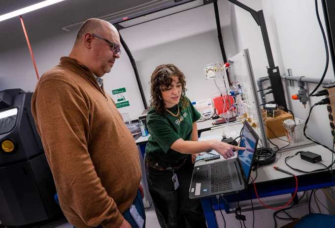Two people work in a laboratory setting. One person is pointing at a laptop screen while the other observes. There are various electronic devices and equipment on the workbench around them.