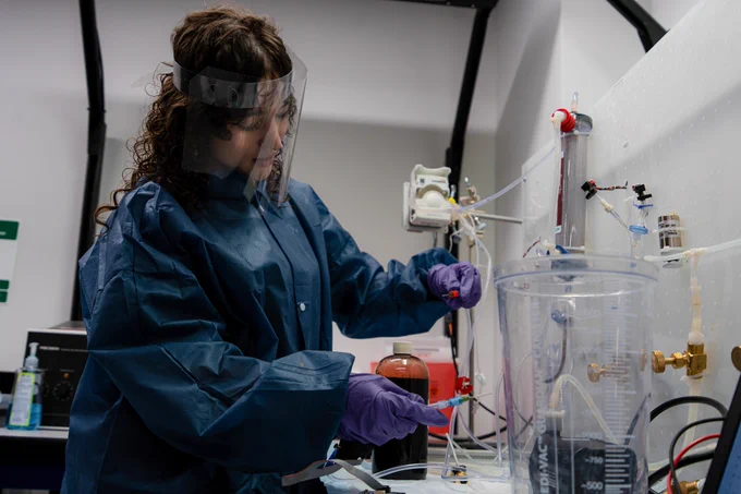 A person in protective gear works in a lab, adjusting equipment on a counter. There are various scientific instruments and containers around them.