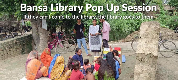 A vibrant outdoor community gathering in a rural Indian village setting for a "Bansa Library Pop-Up Session." In the center, a woman sits on a white floor mat, smiling and engaging with a group of young children and women dressed in colorful traditional sarees. They are gathered around a small, portable wooden bookshelf filled with children's books. Three young adults stand behind the group, observing and participating. The background features lush green trees, bicycles parked against a brick wall, and a glimpse of village life with cattle and thatched-roof structures.