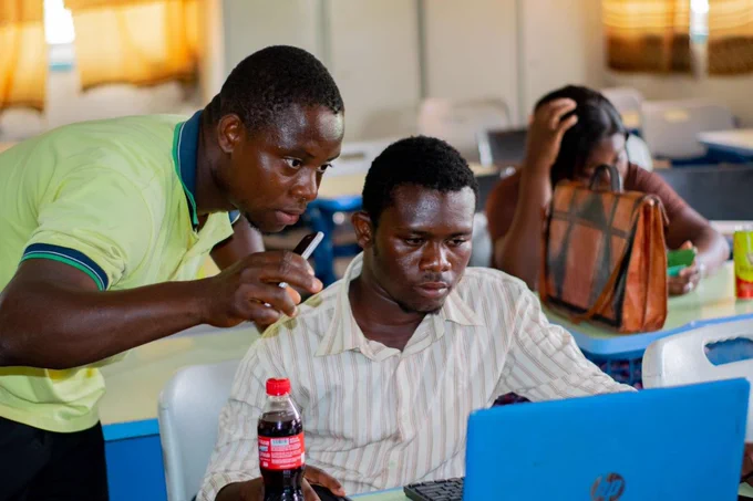 The image shows Isaac Pogser assisting a YouthMappers newbie to make their first OpenStreetMap edit at the UBIDS YOUTHMAPPERS General Meeting on April 11 2026