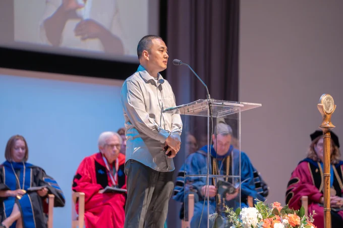Dr. Yi Li, who is wearing a grey button up shit and dark grey pants is standing at a podium introducing Dr. Bleris.
