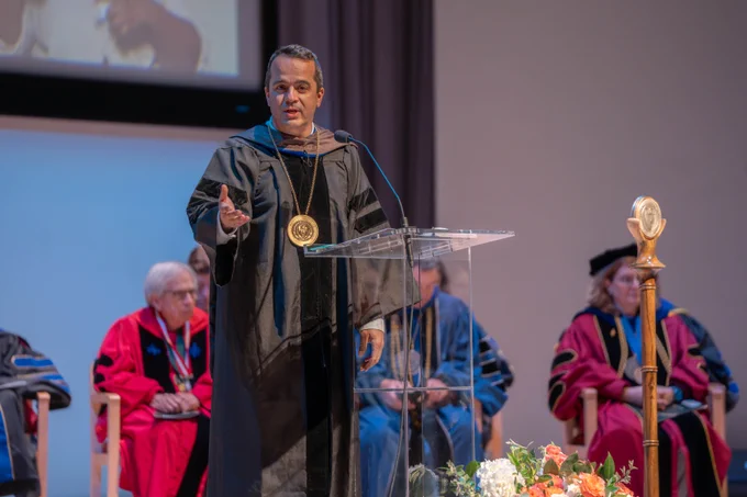 Dr. Bleris is standing at the podium, wearing black regalia, giving a speech.