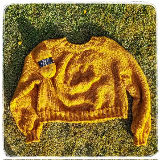 On mossy & dandelion studded garden grass a yellow hand knit jumper is laid out, a ball of wool perched on one shoulder, & little embroidered flowers about the neckline.