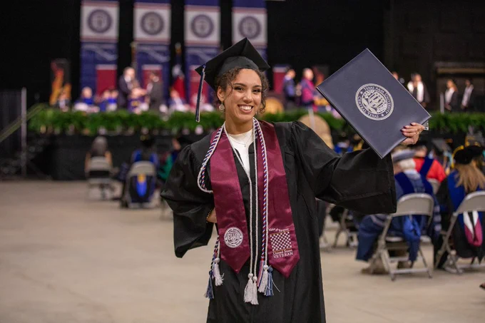 A graduate in a cap and gown holds up a diploma cover, smiling. They are wearing a maroon stole with a logo and standing in an indoor venue with other attendees seated and more graduates and officials on stage in the background.