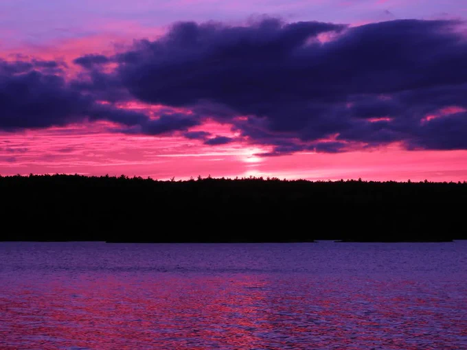 A vibrant sunset over a calm lake, featuring a sky filled with pink, purple, and dark clouds. The silhouette of a forested shoreline is visible in the background.