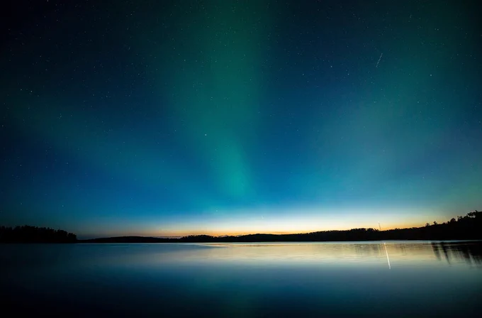 A serene landscape depicts the Northern Lights glowing in green and blue hues over a calm, reflective lake. 