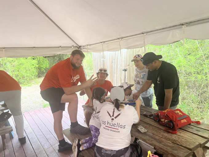 UTRGV football players wearing orange shirts volunteering at Camp Rio in Brownsville, spending time with children and families from the Moody Clinic. Activities include group photos, helping kids into canoes, paddling on the water, and interacting with families under a covered picnic area in a positive, community-focused environment.