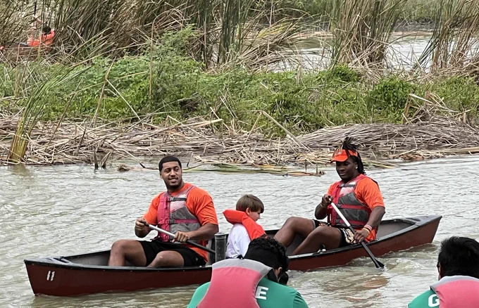 UTRGV football players wearing orange shirts volunteering at Camp Rio in Brownsville, spending time with children and families from the Moody Clinic. Activities include group photos, helping kids into canoes, paddling on the water, and interacting with families under a covered picnic area in a positive, community-focused environment.