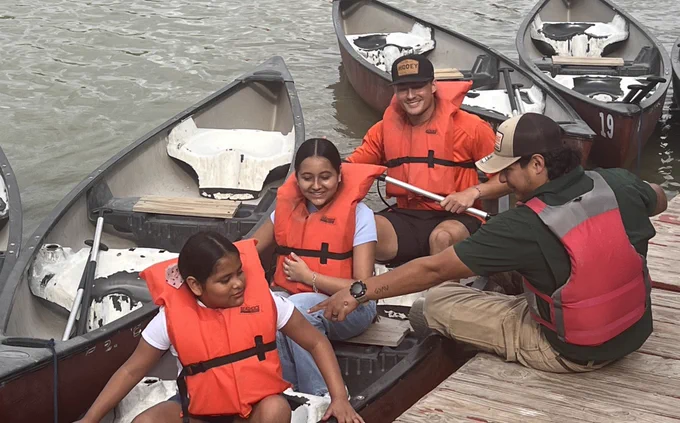 UTRGV football players wearing orange shirts volunteering at Camp Rio in Brownsville, spending time with children and families from the Moody Clinic. Activities include group photos, helping kids into canoes, paddling on the water, and interacting with families under a covered picnic area in a positive, community-focused environment.