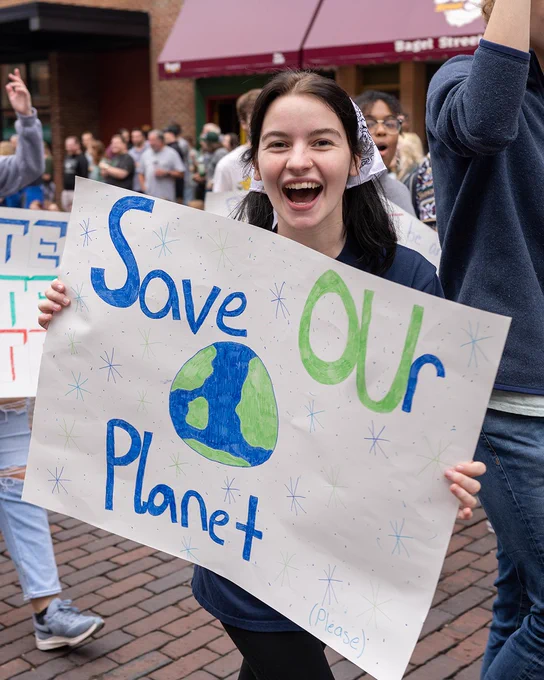 A person is joyfully holding a sign that reads "Save Our Planet" with an illustration of Earth. They are in a crowd at an outdoor event.