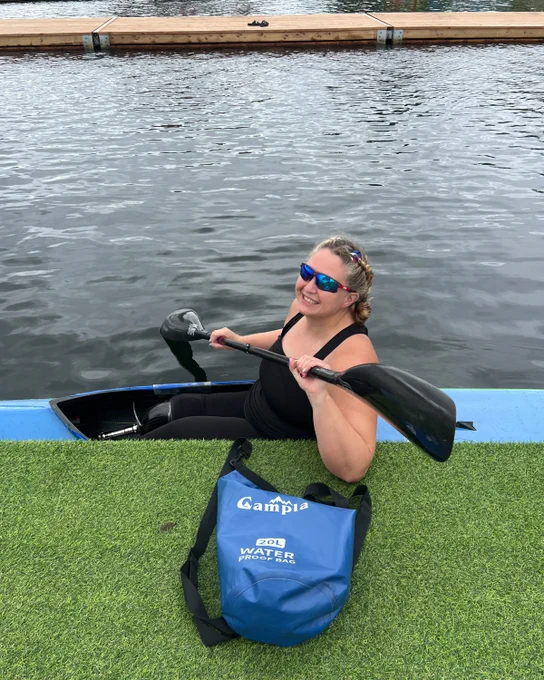 A person in a kayak smiles, holding a paddle. They wear sunglasses. A blue waterproof bag is on the grass beside them.