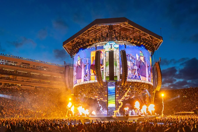 A large concert stage at Notre Dame Stadium is illuminated with bright lights and surrounded by flame effects. Huge screens display performers and vibrant visuals. The stadium is filled with a large, energetic crowd under a darkening evening sky.