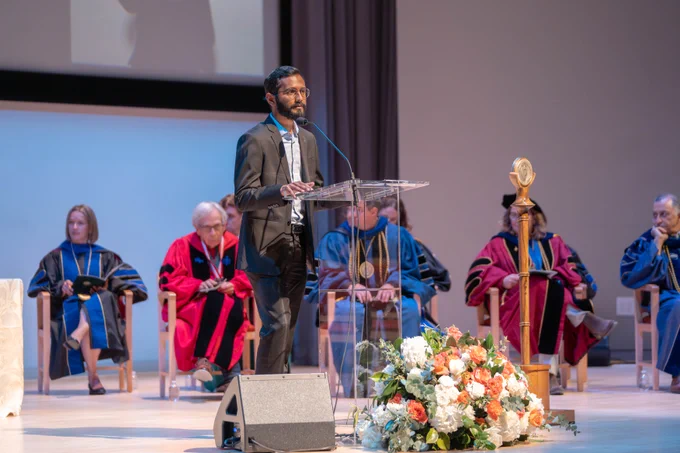 Dr. Nimit Shah is at a podium wearing a black suit and a grey shirt, introducing Dr. Obaid. Faculty in the background are wearing regalia and there are white and orange flowers in front of the podium