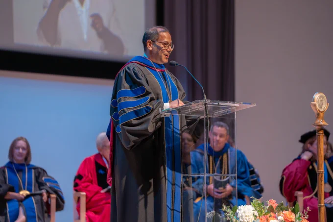 Dr. Obaid is standing at a podium wearing black and blue regalia, delivering a speech