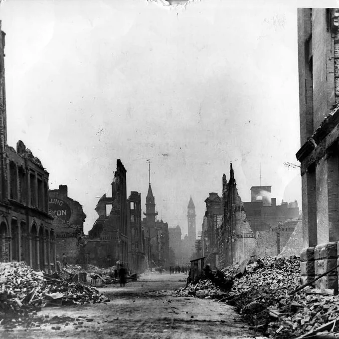 Black-and-white photograph of a wide Toronto street in 1904, lined with heavily damaged buildings on both sides. Brick facades are partially collapsed, with exposed interiors, broken walls, and empty window openings. Large piles of rubble and debris fill the sidewalks and spill into the roadway. In the distance, several small figures stand or walk along the street. A clock tower and other taller structures rise faintly through the haze in the background. The foreground shows uneven ground covered with dust and scattered debris.
