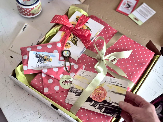 Verpacke eine Bestellung in rotem Geschenkpapier mit weißen Punkten und mit Schleife, diverse Extras liegen bei, und von der Seite reicht meine Hand eine Karte mit Rosenmotiv und der Aufschrift danke dazu 