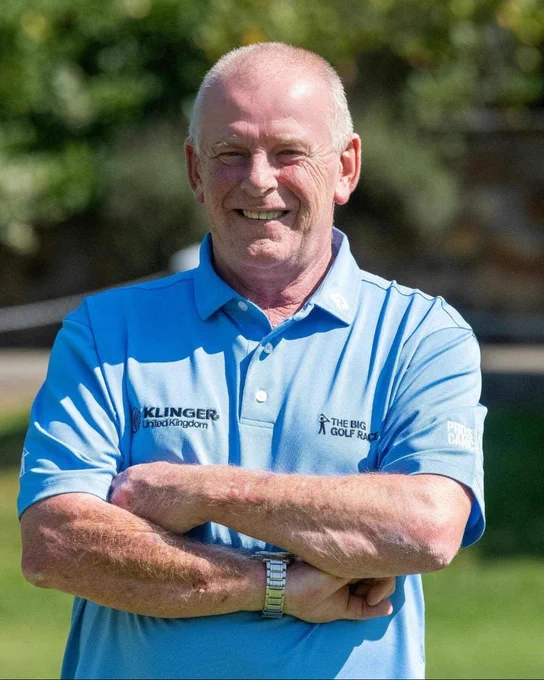 Jim Finnie stands with his arms crossed, smiling at the camera. He wears a The Big Golf Race polo shirt. In the background is a golf course.