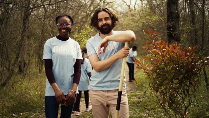 A smiling man and woman wearing light blue "volunteer" T-shirts stand in a wooded area. 
