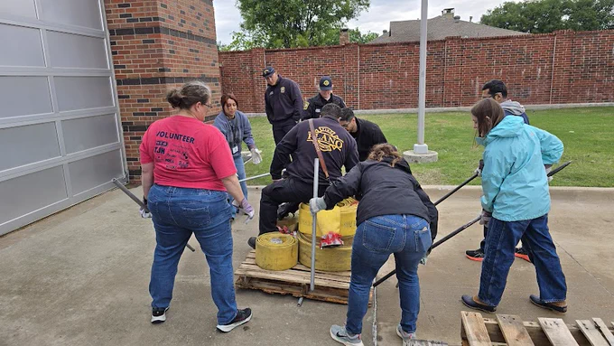 A group of people outdoors, working together to lift a heavy weight using long metal rods and wooden pallets. They are dressed in casual clothing with some wearing gloves. A brick wall and grass are visible in the background.