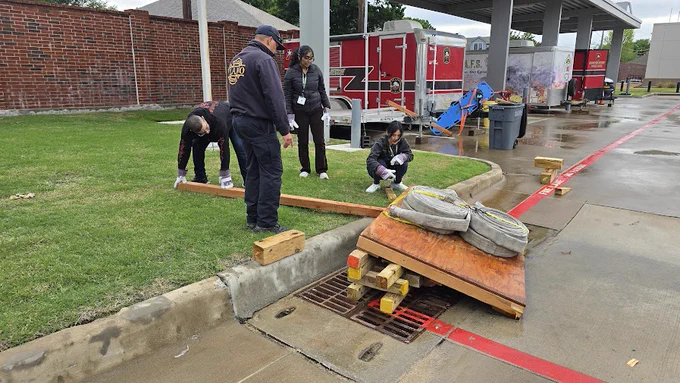 Four people are participating in a flood simulation exercise near a curbside storm drain. They are setting up a wooden ramp with hoses across the drain. Two individuals are kneeling on the grass, while two others stand and observe. Behind them is a wet sidewalk, red trailer, and emergency equipment.