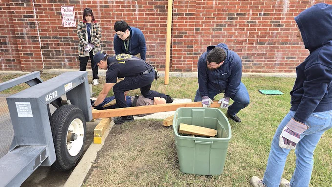 A group of people are gathered around a mechanical trailer with blocks of wood. One person is kneeling and reaching under the trailer, while another assists by holding a piece of wood. A large green bin containing more wood pieces is nearby. The scene is outdoors on grass, next to a brick wall with a "Facilities Maintenance Parking Only" sign. Some individuals are wearing gloves and jackets, suggesting cool weather.