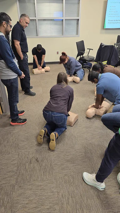A group of people are practicing CPR on mannequins in a classroom setting. Several individuals are kneeling on the carpeted floor, focused on performing chest compressions. A man stands nearby, observing and providing guidance. Chairs and a projector screen with text are in the background.