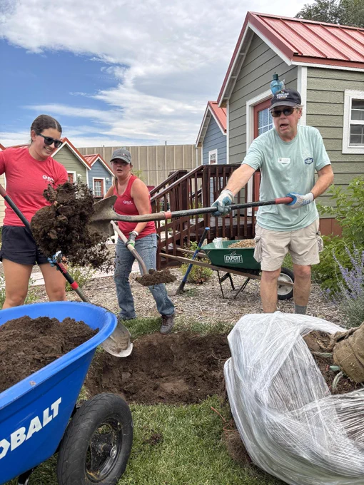 Three people are actively gardening, with two of them using shovels to move soil towards a wheelbarrow labeled "Cobalt." They are standing in front of small houses with red roofs. A large wrapped plant is on the ground nearby. The scene takes place outdoors on a grassy area under a partly cloudy sky.
