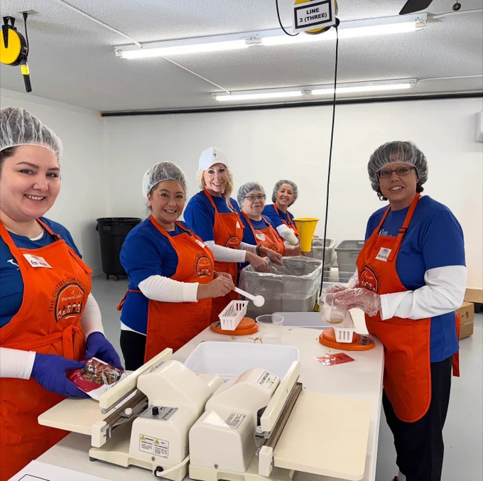 A group of people wearing blue shirts, orange aprons, hairnets, and gloves are standing around a table in a bright room, packing food items. They are smiling and appear to be volunteering.